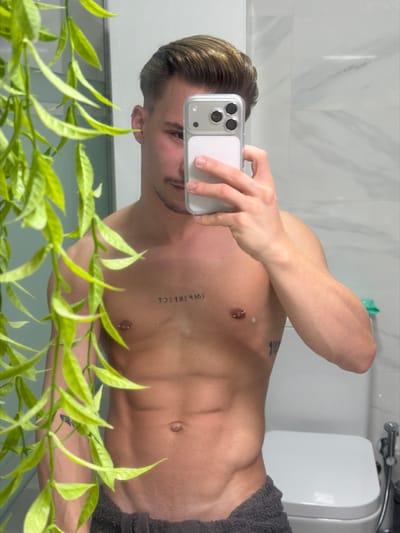 A male individual is depicted in a mirror selfie taken within a modern bathroom located in Viseu, Portugal. The subject, shirtless, possesses a muscular physique with defined abdominal muscles. He has styled brown hair with lighter highlights and exhibits facial hair consisting of a mustache and stubble. A piercing is visible in his left ear. Two barbell piercings are present on his nipples, and a tattoo reading "IMPERFECT" (reversed in the reflection) is visible on his upper chest. A second, partially obscured tattoo is present on his left side. He is shown holding a light-colored smartphone with a clear case and triple camera lenses in his right hand, which partially covers his face as he photographs his reflection. The lower portion of his body is covered by a dark grey towel wrapped around his waist. The bathroom environment features white, rectangular wall tiles with subtle grey veining. A white, modern toilet with a closed lid and a chrome bidet sprayer are visible in the background reflection to the subject's right. A green, leafy plant hangs into the frame from the left, partially obscuring the scene.