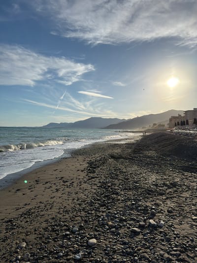 This media file shows a view of a beach in Vallecrosia al Mare, Italy. The image is oriented vertically, with the sky occupying the left side and the beach, water, and a distant coastline on the right.
The sky is a vibrant blue with wispy white clouds and a bright sun, suggesting it is daytime, likely late afternoon or early morning due to the warm light and long shadows. An airplane can be seen in the upper left portion of the sky, leaving a contrail.
The beach itself is composed of dark sand and numerous smooth, gray pebbles. The sea is a clear, greenish-blue, with gentle waves lapping at the shore. In the distance, mountains rise, with a town or buildings visible nestled at their base, partially obscured by haze or the angle of the sun. A few dark umbrellas are set up on the beach in front of the buildings, indicating a possible beach resort area.
Two figures are walking along the water's edge in the mid-ground. The overall mood is serene and tranquil, evoking a sense of a peaceful seaside setting. There is no visible text in the image.