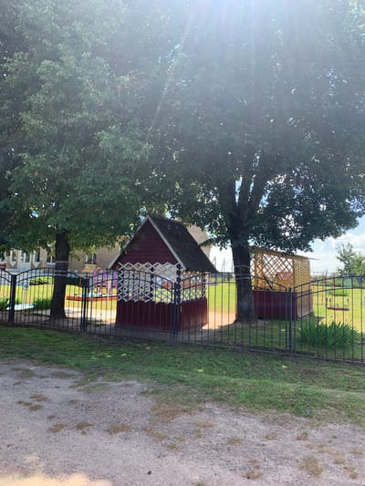 The image shows an outdoor playground area with two small wooden structures, a fence, and trees. The larger structure, on the left, is a red, hexagonal gazebo with white lattice work on its sides and a dark roof. To the right is another, larger wooden structure, a yellow, octagonal gazebo with similar lattice work. A swing set is visible behind the yellow gazebo. The playground is enclosed by a black metal fence. In the background, a building with a light-colored brick facade is visible, suggesting a school or community center. The ground is a mix of gravel and grass. The image is taken on a sunny day, with strong sunlight filtering through the leaves of the large trees. This scene is located in the vicinity of Слабада-Кучынскі сельскі Савет, Belarus.