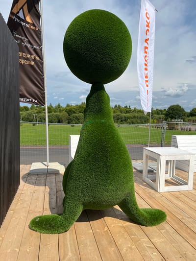In Yakunino, Russia, a green, grass-covered sculpture resembling a seal balancing a large sphere on its head stands on a wooden deck. The sculpture is positioned near a metal fence that encloses a grassy area with trees in the background. Two flags are visible: one dark brown with orange text and a logo, and a white flag with orange text. White wooden benches and a table are placed on the deck to the right of the sculpture. The sky above is partly cloudy.