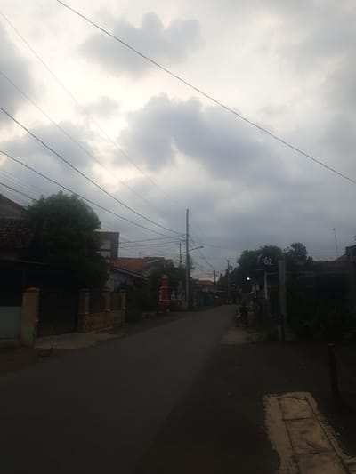 The image depicts a street scene in Proyonangg, Indonesia, under an overcast sky. The street is paved and appears to be residential, with houses and fences lining its sides. Overhead, a dense network of electrical wires crisscrosses the frame. On the left side of the street, a house with a tiled roof and a fence is visible, partially obscured by a large tree. On the right side, a sign with "+62" is mounted on a pole, and a person is standing next to a bicycle. The overall impression is one of a typical suburban or semi-urban environment in Indonesia, with everyday infrastructure and a muted, cloudy atmosphere.
