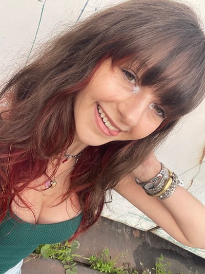 A young woman smiles and sticks out her tongue in a selfie. She has brown hair with red highlights and bangs. Her skin has a light tone and she appears to be wearing white eyeshadow. She wears a green tank top and multiple bracelets on her left wrist. She also wears several necklaces. A white wall with blue paint streaks is to the left of her and a concrete ledge with weeds is on her right.