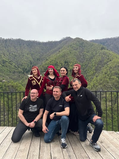 On a wooden deck overlooking a green, mountainous landscape in Kveda Chkhutuneti, Georgia, a group of six individuals are posing for a photo. Four women are wearing traditional Georgian attire of maroon and gold, including head coverings. They are arranged vertically on the left side of the image. Three men are arranged vertically on the right side. They are wearing dark-colored casual clothing. There is a metal railing between the group and the background scenery. The sky is overcast.