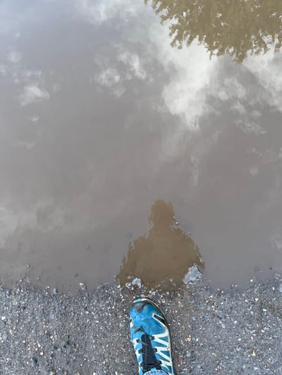 A top-down view features a muddy puddle reflecting the sky with scattered clouds and the silhouette of a tree. Adjacent to the water's edge is a rough, gravelly surface. A blue and black athletic shoe sits partially on the gravel and slightly overlaps the puddle. The overall composition is dominated by the contrasts between the reflective water, the rugged ground, and the colorful shoe.
