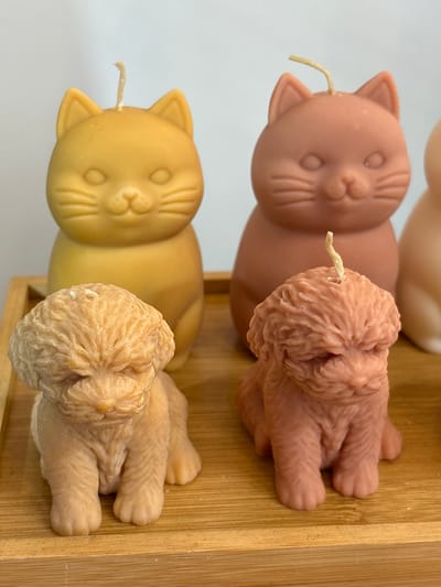 The image shows two animal-shaped candles in a light-colored wooden box. The candles are shaped like a cat head connected to a dog body. The top candle is brownish-pink, while the bottom candle is yellow. Both candles have a wick protruding from the point where the cat and dog parts meet. The background is a white wall. There is no evidence in this image to suggest a location in the city of Chorrillos, Peru.