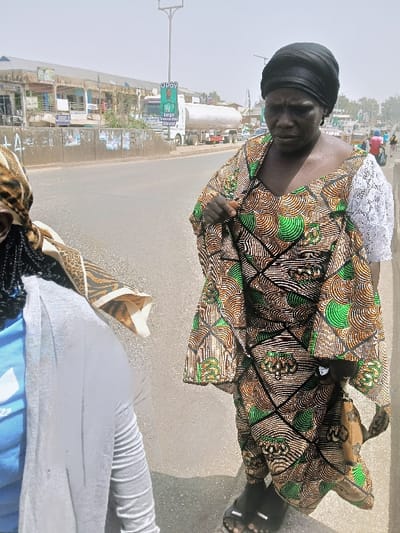 An urban street scene in Kadoma, Zimbabwe, features two individuals positioned in the foreground. On the right, an adult female with a dark complexion wears a black headwrap, a brown, green, and black patterned dress with white lace sleeves, and black open-toed sandals. A small, patterned brown purse hangs from her left side. Her head is tilted downwards, and her expression appears solemn. In the left foreground, the back of another individual is partially visible, wearing a patterned headscarf, a light blue shirt, and a light grey cardigan, with a piece of patterned fabric draped over their left shoulder. The background reveals a paved road with a concrete median adorned with various posters. A white tanker truck is visible on the road, and an "OPay" sign on a tall pole is positioned further back. Commercial buildings line the street, indicating an active urban area. The sky is bright and hazy, consistent with a daytime environment.