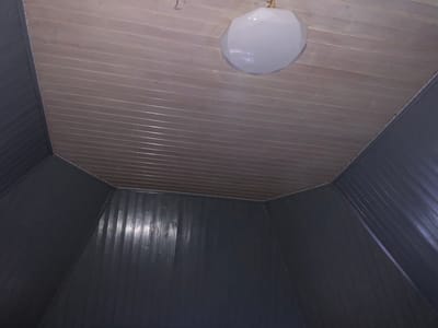 The image shows the interior of a room with paneled walls and a paneled ceiling. The walls are dark gray and have a vertical paneling pattern. The ceiling is made of lighter colored wood planks that run horizontally. A white, polygonal light fixture is attached to the ceiling. The room appears to be somewhat angular. There are no people or animals visible in the image. The lighting is dim, with the light fixture being the primary source of illumination. The scene is likely a residential interior.