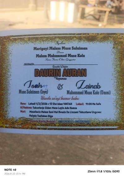 A rectangular paper document, identified as a wedding invitation ("DAURIN AURAN"), is depicted in a slightly angled, close-up shot against an out-of-focus background. The document details the marriage ceremony between Isah Musa Sulaiman and Zainab Muhammad Musa Kofa, scheduled for Sunday, February 1, 2026 (13 Sha'aban 1447 AH) at 11:00 AM. The gathering point is specified as Tukuntawa Gidan Maza Layin Ado Hamza. The main venue for the ceremony is the Malam Sani Mosque, located in Mai Kwado Da Linzami, within the Tukuntawa Unguwar Malado Tsallaken Diga area, which is situated in the city of Tudun Wazurchi, Nigeria. The document features a white background with decorative golden-brown borders and black text. Two contact phone numbers (08032938775 and 08032828837) are listed for inquiries. The background is blurred, showing light-colored ground and a faint, indistinct wooden structure in the upper right, suggesting an outdoor or brightly lit indoor setting. No individuals are visible.
