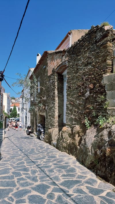 The image depicts a narrow, cobblestone street in Cadaqués, Spain. A steep, rocky hillside borders the right side of the street, with a rough stone wall constructed at its base. Several buildings with red-tiled roofs and whitewashed facades line the left side of the street. Some of these buildings appear to be old, with visible brickwork and rough stone construction elements.
A few people are visible walking down the street, seemingly engaged in casual conversation. Among them are men, women, and children, dressed in summer attire. A black motorcycle is parked on the left side of the street, partially obscured by a tree with sparse foliage. Another motorcycle is parked further down the street, also on the left.
The sky is a vibrant, clear blue, suggesting a sunny day. The lighting indicates it is likely midday, with distinct shadows cast across the cobblestone pavement by the buildings and a thin wire stretching across the scene. The overall impression is a charming, sun-drenched European village street. There is no visible text in the image.