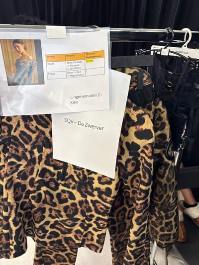 The image captures a detailed view of a backstage area at a fashion event in Herentals, Belgium. A black metal clothing rack dominates the scene, from which various garments and paper documents are suspended.

In the foreground, a prominent leopard print athletic-style top, featuring a black zipper and partially revealing a "GUCCI" brand inscription near the neckline, hangs on the rack. To its right, several black lingerie items are displayed on white plastic hangers, one of which is branded "CHANTELLE." These items include what appears to be a black corset or bustier with gold-colored eyelets and lace detailing.

Attached to the clothing rack are multiple paper documents. One document, enclosed in a transparent plastic sleeve, displays a photograph of a female model wearing a blue garment with intricate embellishments. Adjacent to this photograph, a table with a schedule is presented in Dutch, detailing "Timing," "Wereld" (World), and "Tijd voor kledingwissel" (Time for costume change). Entries include "20u48" for "Show 1B - EQV" with an "18 MIN" costume change allowance, and "21u09" for "Show 7 - GLE." Text beneath the table identifies "Lingeriemodel 2 : Kiko." Another partially visible paper below reads "EQV - De Zwerver."

In the lower right quadrant of the image, the lower legs and feet of an individual are visible, clad in tan, fur-lined slippers. The background consists of a dark, likely black, fabric curtain or drape, indicative of a temporary event space.