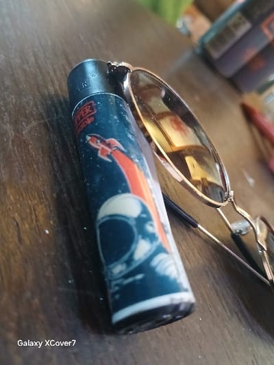The scene depicts two main objects resting on a reddish-brown wooden surface with visible grain. A dark-colored cylindrical lighter, identified as a "Clipper Refillable" brand, is positioned prominently. Its casing features a space-themed graphic: an orange rocket with a red contrail ascending, and a stylized white and dark blue astronaut helmet. The top of the lighter is black and shows a faint registered trademark symbol. To the right of the lighter, a pair of sunglasses with metallic, possibly gold-toned, frames and dark, reflective lenses is partially visible. The left temple arm of the sunglasses extends to the right. Reflections in the sunglass lenses show blurred interior details. The background is out of focus, revealing indistinct shapes that appear to be other objects, possibly containers, and a red elongated item. No individuals are present in the image. The image itself provides no visual information to determine a specific location within Montabaur, Germany. A "Galaxy XCover7" watermark is present in the lower-left corner of the image.