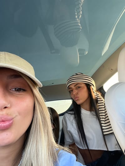 The image depicts three individuals inside a modern vehicle with a panoramic glass roof, located in Jaljulia, Israel. In the foreground, positioned to the left, is a woman with blonde hair, wearing a light-colored baseball cap and a light blue top. A silver nose ring is visible on her left nostril, and her lips are pursed forward. To her right, in the mid-ground, is another woman with long dark hair, wearing a black and white striped head covering that extends over her shoulder. She is dressed in a white cropped t-shirt and dark lower garment, with her lips also pursed forward. A third individual with dark hair is partially visible in the background between the two primary subjects. The car's interior features white seats. The glass roof reflects an inverted image of the woman with the striped head covering and portions of the car's interior. Sunlight enters from the right side of the frame, indicating an outdoor setting, though the exterior view is blurred. All visible individuals appear to be looking towards the camera.