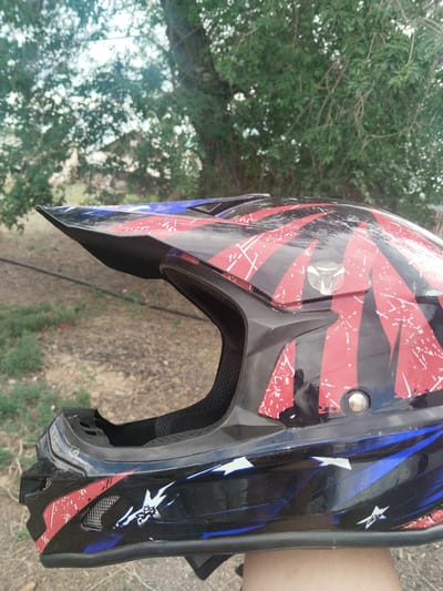 This is a close-up, medium shot of a black and red motocross helmet with blue accents and white star graphics. The helmet is angled to the right, showing its profile. The sun visor is prominent and angled upwards. The interior padding is visible through the opening. The helmet has a rugged, worn appearance with some scuffs and scratches. In the background, there is out-of-focus foliage from trees and dry, earthy ground, suggesting an outdoor setting in a natural environment. The lighting indicates it is daytime. The image was taken in Ақадыр, Kazakhstan. No text is visible.