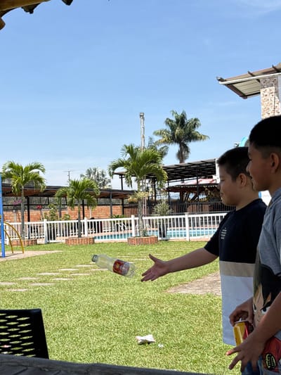 The scene depicts an outdoor recreational area in Pitalito, Colombia, under a clear blue sky. In the foreground, a dark, textured surface is partially visible. Two young males are positioned on the right side of the frame. The male closer to the center, wearing a blue, white, and tan striped shirt, has his right arm extended with his hand open, palm upward. Directly to the left of his hand, a clear plastic bottle, partially filled with a yellow liquid and featuring a red and white label, is suspended in mid-air in a horizontal trajectory. A second male, partially visible on the far right in a grey t-shirt with a graphic design, holds two beverage cans and is observing the action. On the green grass beneath the bottle, a small piece of crumpled white and red litter is present. The midground features a manicured green lawn with light-colored stepping stones. Beyond the lawn, a white picket fence encloses a blue swimming pool and several shaded structures with dark metal frames and roofs. Multiple palm trees are scattered across the background, and a tall metal pole topped with a light fixture is visible in the distance.