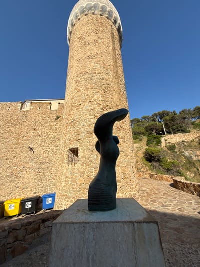 The image shows an outdoor scene with a tall, stone tower in the background and a dark abstract sculpture in the foreground. The tower has a mesh-like covering on its top. To the left of the tower, there is a stone wall and three recycling bins (yellow, black, and blue). The ground is paved with stones. The environment appears to be a sunny day with a clear blue sky. The scene is located in the vicinity of the Vila Vella, the old medieval town of Tossa de Mar, Spain, which is a fortified old town on the coast. The sculpture is positioned on a stone pedestal in front of the tower and a section of the town wall. A path paved with irregular stones leads away from the sculpture and towards the hillside, which is covered in trees.