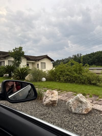 The image shows a view from inside a car, likely a passenger side window. Reflected in the side mirror are a person's hands holding a cell phone. Outside the car, the scene is dominated by a two-story, white stucco building with a dark brown roof. The building is surrounded by landscaping, including various green plants, bushes, and trees. There are large, light-colored rocks in the foreground near a gravel area. Overhead are power lines against a cloudy sky. While the city, Jinhua, is provided, there are no definitive elements within the image to confirm the exact location.