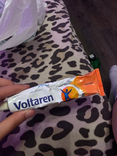 A tube of Voltaren, an anti-inflammatory gel, is being held by a person's hand against a leopard-print fabric. The tube is white and orange, with the brand name "Voltaren" prominently displayed in blue lettering. The text on the tube is in Arabic and appears to describe the product's uses. The background is a dark wood-like surface. A white plastic bag is partially visible in the upper left corner of the image. The image was taken in Vaganovo, Russia.