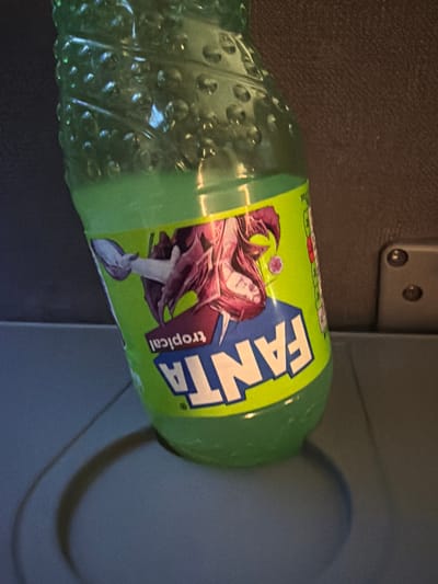 The image shows a green plastic bottle of Fanta Tropical beverage, lying on its side. The bottle has a textured surface with raised bumps. The label on the bottle features the Fanta logo in blue and white, with the word "Fanta" prominently displayed. Below the logo, there is an illustration of a person with long, dark hair and red clothing, possibly a character from a fantasy or anime-style artwork. The word "tropical" is printed vertically in red text next to the illustration. The bottle is positioned in what appears to be a dimly lit interior, possibly a vehicle or an indoor setting with limited natural light, suggested by the shadow cast on the right side of the image. The background on the left is a dark, textured surface, possibly upholstery or fabric. On the top left, a metallic fixture with two screws is visible, which could be part of a seat mechanism or interior fitting. The overall lighting is subdued, with the Fanta bottle being the primary illuminated subject.