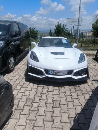 The image shows a white Corvette with license plate "RA-021-SL" parked in a lot paved with interlocking concrete blocks. A black van is parked to the left of the Corvette. A billboard is visible in the background and shows a Georgian phone number, indicating the location is Georgia. The image was taken in the city of Rustavi, Georgia.