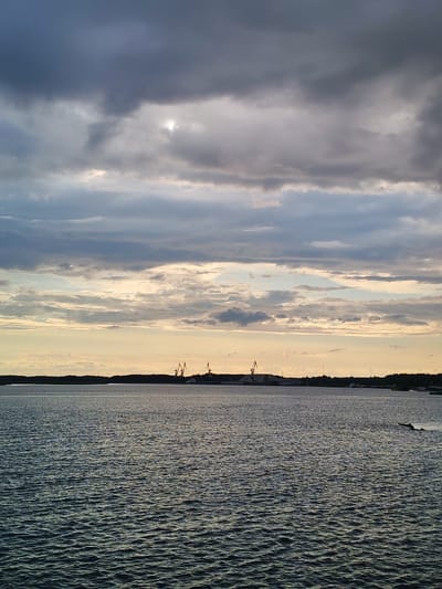 The image depicts a serene body of water under a dramatic sky filled with dark, textured clouds. A sliver of lighter sky with pale sunlight breaks through the clouds near the center, casting a soft glow. The water's surface is rippled, reflecting the subdued light. In the distance, a dark, tree-lined horizon separates the water from the sky. Silhouetted against this horizon are several tall industrial cranes, suggesting a port or industrial area. The scene appears to be a coastal view within the city of Shalsky, Russia, likely captured during the late afternoon or early evening, given the lighting and cloud formations. There are no visible people or animals in the frame.