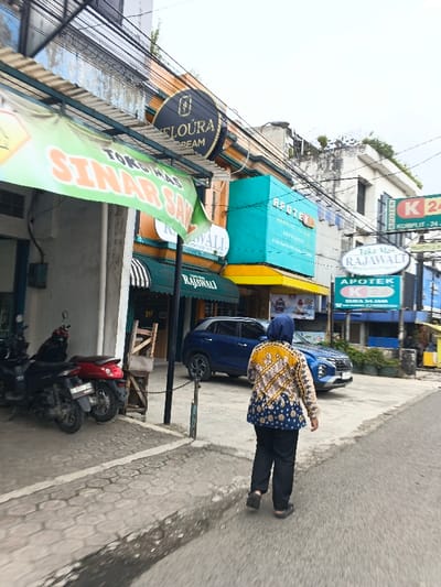 The image captures a street scene featuring a woman walking away from the camera. She is wearing a patterned yellow and blue top with dark pants and a headscarf. Various storefronts line the street with visible signage like "TOKO MAS SINAR SAK," "FLOURA CREAM," "APOTEK," and "RAJAWALI."  A blue SUV is parked on the side of the road. A few motorcycles parked on the sidewalk add to the urban environment. Overhead, a tangle of wires suggests a busy thoroughfare.