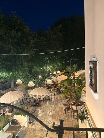 This is an outdoor nighttime scene at a restaurant located in Barletta, Italy. The courtyard of the restaurant is paved with stone tiles and features several tables and chairs for outdoor dining. Large, white, fringed umbrellas provide shade. Woven pendant lights hang from trees above, and string lights are also visible, casting a warm glow over the area. Lush greenery, including large palm-like plants and trees, surrounds the courtyard. In the background, a car is parked on a street. A building with a light-colored facade and a window with wooden shutters is visible on the right side of the frame. A few patrons are seated at tables, engaged in dining or conversation. The overall atmosphere is that of a relaxed, open-air dining establishment.