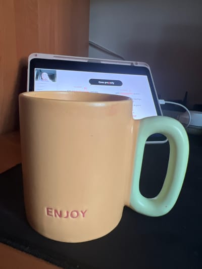 The image shows a light orange mug with the word "ENJOY" embossed in pink on its side. The mug has a light green handle and is placed on a black surface in front of a tablet device. The tablet screen displays a video call with a young woman's face in a small window at the top left corner, and text in various languages, including Polish and Russian, beneath it. The background appears to be an indoor setting with wooden furniture on the left. The scene is brightly lit.