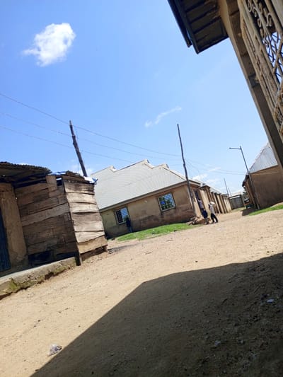 The image depicts a sun-drenched, dusty street in Barakin Babele, Nigeria. Two children are visible in the middle ground. One child, wearing a light-colored shirt, is carrying a large white bucket, suggesting they are collecting water or performing some chore. The other child, wearing a darker shirt, stands nearby. The street is unpaved, with loose sand and gravel.
On the left side of the image, a makeshift structure is visible, constructed from rough wooden planks and what appears to be mud or clay. It has a corrugated metal roof. Adjacent to this structure is a more substantial building with a light-colored, textured wall and a corrugated metal roof. This building has a barred window. Further down the street, other buildings with similar corrugated metal roofs and textured walls are visible, indicating a residential area. A street light stands to the right of the main building. A small, three-wheeled vehicle, possibly a motorcycle taxi or tuk-tuk, is parked further down the street. Overhead, power lines crisscross the sky, supported by wooden poles. The sky is a clear blue with a few scattered white clouds. The overall impression is one of a typical, informal urban environment in Nigeria.