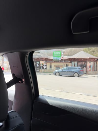 The image depicts an interior view from a car in Gintsi, Bulgaria. The car's sunroof is open, revealing a street scene. A parked light blue car is visible outside. The street has buildings on the side. The car's interior includes the headrest of a seat, a seatbelt attachment, and the dark-colored roof and panels of the car. The environment is typical of a small Bulgarian town. There are no people visible.
