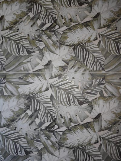 This is a close-up, overhead shot of a wall with a tropical leaf pattern. The leaves are rendered in shades of green, gray, and white, with some appearing more olive green, while others are a pale, desaturated green. The pattern features large, broad leaves with visible veins and some with characteristic fenestrations, suggesting they are monstera leaves, alongside palm-like leaves with segmented fronds. The style is painterly or possibly a textured print, with a subtle sheen on some of the lighter areas.

A thin, horizontal line, possibly a grout line or a seam, divides the image roughly in the middle. The lighting appears to be even and diffused, with no distinct shadows or highlights that would suggest a specific time of day or natural light source. The setting is a wall, likely indoors, given the texture and finish of the material. There is no discernible activity or event taking place, as the image focuses solely on the static pattern. No people, animals, or any other objects are visible. The overall mood is neutral, focusing on the aesthetic of the decorative pattern. There is no visible text in the image. The location context of Trudobelikovskiy, Russia, does not provide any visual cues within the image itself.