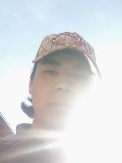 The image depicts a single individual, reportedly located in Takhtakuprik, Uzbekistan. The subject, an adult male with discernible East Asian or Southeast Asian features, is presented in an outdoor setting under intense natural light. He is wearing a beige baseball cap featuring a repeated "GG" monogram pattern and a red and black king snake design, consistent with a Gucci branded item. A dark, zippered garment is partially visible on the lower left portion of the image. A powerful light source, presumed to be the sun, is positioned behind the subject, causing significant backlighting, severe overexposure, and lens flare that obscures the right side of the subject's face. The visible background consists primarily of a light blue sky, with a faint circular object discernible in the upper left quadrant. A dark, indistinct structure with a sloped roofline is partially visible in the far left background.