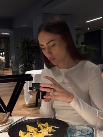 The image shows a female individual seated at a table in a modern restaurant located in Torbole, Italy. The subject, possessing long brown hair and wearing a white long-sleeved top with a silver necklace and multiple rings on her left hand, is actively holding a black compact digital camera with both hands, aiming it downwards. Positioned directly in front of her is a black textured plate containing yellow pasta shapes garnished with a white sauce and dark shavings, presumably truffles. To the left, a slice of bread rests on a small white plate, and a clear glass with a distinct teal swirl pattern is visible on the right. The table is covered with a white tablecloth. The background features a restaurant interior characterized by dark walls, a light wooden floor, potted green plants, additional dining tables with white coverings, and modern lighting fixtures, including recessed and strip lights. Other patrons are faintly discernible in the blurred distance.