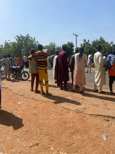 This image depicts a daytime scene in Lago, Nigeria, under a clear blue sky with a high sun angle casting prominent shadows. A gathering of multiple individuals, predominantly male, stands along the edge of a dark asphalt road.

The foreground consists of dry, reddish-brown earth containing sparse plant debris and a single transparent plastic wrapper. On the left mid-ground, a dark-colored motorcycle with spoked wheels is parked, facing right, surrounded by several standing individuals. One individual in an orange safety vest is visible further back in this area.

A line of men occupies the center and right mid-ground, positioned along the edge of the asphalt road and mostly facing away from the camera. Two prominent figures on the left of this line include one man in a horizontally striped yellow, red, and black long-sleeved shirt and yellow pants, with his arm around another man dressed in a light-colored top and dark red pants. Other men in the line wear traditional Nigerian garments, including long robes (agbada) in colors such as maroon, cream, and light pinkish-brown, some with matching caps. One individual features long, dark braided hair.

The background reveals a dense line of green leafy trees. A utility pole with multiple power lines extends vertically on the right side against the clear sky. A grey-roofed building is partially visible behind trees on the far left. Individuals in the scene are primarily static, standing and observing the general area.