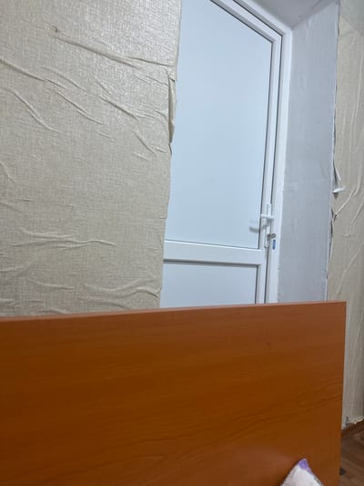 In Corlăteni, Moldova, a close-up shot focuses on a white door with a silver handle and a small key attached, set against a wall with peeling beige wallpaper. The bottom of the frame is dominated by a wooden surface, likely a desk or bed, with a visible wood grain pattern.