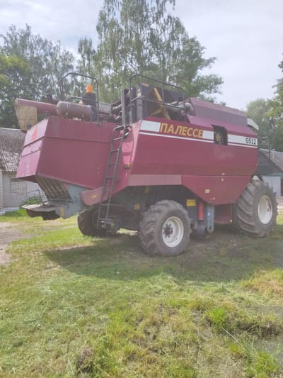 A red and white combine harvester, identified by the markings "ПАЛЕССЕ" and "GS12", is parked on a grassy area with some cut grass and a dirt path. The combine has large agricultural tires and a ladder on its side. In the background, there is a white brick building with a grey roof and a line of trees. The sky is cloudy. The image appears to have been taken in an rural setting, possibly in or near Залескі сельскі Савет, Belarus, given the context. There are no people visible in the image.