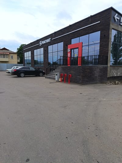 This is a medium-angle exterior shot of a Magnit supermarket in Kasumkent, Russia. The building has a modern design with a dark brick facade and large, reflective windows. A bright red entrance archway is visible, with the Magnit logo prominently displayed above the windows in white lettering. The time of day appears to be late afternoon or early evening, with overcast skies suggesting a cloudy or slightly rainy day.
In the foreground, a grey asphalt parking lot occupies most of the frame. Two cars are parked on the left side of the lot: a dark sedan and a lighter-colored sedan behind it. To the right of the building, a set of stairs leads up to the entrance. Red bollards are placed along the sidewalk in front of the building.
Notable details include the clear signage of the Magnit store and the architectural style of the building, which appears to be a contemporary retail space. The overall impression is one of a functional, modern commercial establishment in a town or suburban setting. No people are visible in the image.