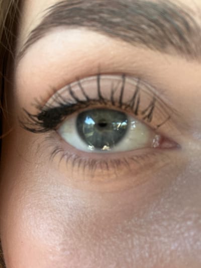 A close-up, eye-level, medium shot captures a person's left eye, illuminated by what appears to be a natural light source reflecting in the pupil. The eye itself is a blue-green hue, framed by dark, applied mascara on the upper lashes and a lighter, more sparse application on the lower lashes. The skin around the eye is fair with some visible texture and what appear to be small white blemishes or milia below the lower lash line. An eyebrow, filled and shaped with dark product, arches above the eye. The background is out of focus, but suggests a neutral, indoor environment. The image does not contain any information or context that would allow for its location to be determined in Everett, United States of America.