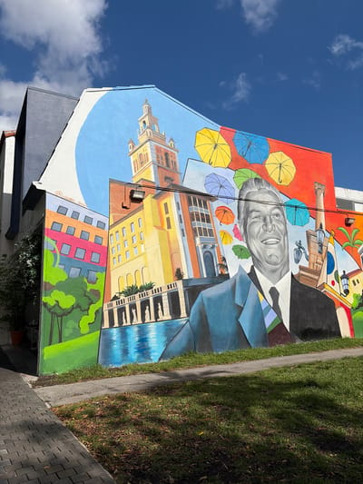 The image depicts a mural on the side of a building in Coral Gables, Florida, United States of America. The mural features a portrait of a smiling man in a suit and tie, surrounded by colorful umbrellas and a rendering of the city's iconic architecture, including the Coral Gables City Hall. Below the City Hall, there is a building. The mural extends down to street level and is adjacent to a sidewalk and grassy area. A signature is visible in white text near the bottom of the mural. The sky above is blue with some clouds.