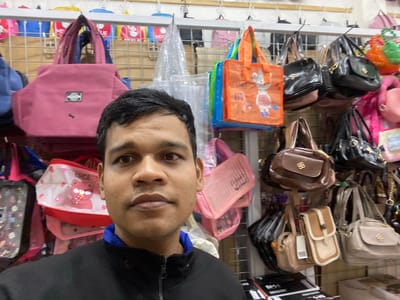 This media file shows a person, likely a man, taking a selfie in what appears to be a retail store selling bags and accessories. The camera is positioned low, capturing the lower half of his face, including his eyes, nose, and mouth, along with his dark hair and a black jacket with a blue lining. Behind him, shelves are filled with various bags of different sizes, colors, and materials. These include light-colored crossbody bags, brown leather bags, pink pouches, and orange reusable shopping bags with cartoon prints. Some of the bags have visible branding or decorative elements. The setting suggests an indoor market or a shop specializing in handbags and personal accessories. The lighting is artificial, typical of an indoor retail environment. The person's expression is neutral, and the overall mood is casual. The image doesn't provide clear cues about the time of day or weather. Visible text includes "BMW" on black packaging in the background and some text on the orange bags that is difficult to decipher. Given the location context of Lamteumen Barat, Indonesia, the scene likely takes place in a shop there.