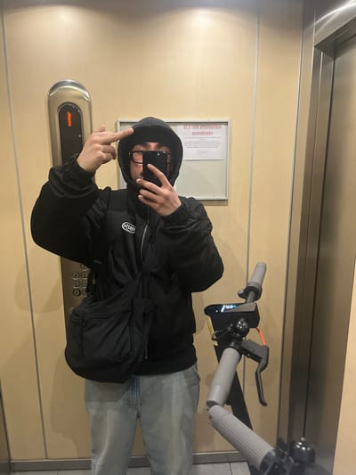 A person wearing a black hoodie, glasses, and light-wash jeans is taking a selfie in an elevator. The person is extending their middle finger towards the elevator button panel. Next to the person is an electric scooter. The elevator interior has beige walls and stainless steel trim. There is a framed notice posted on the wall. The location is Aubervilliers, France.