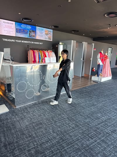 A young man wearing glasses, a black shirt, and black pants walks past a mirrored counter decorated with white circular patterns. He is holding a mobile phone in his right hand. In the background, a rack of colorful traditional Vietnamese Ao Dai dresses is visible, along with mannequins dressed in red and pink garments. Above the counter, a digital display shows information about "Ao Dai & Hanbok for Rent," "Unique Reality Games," and "Photo Service." The environment appears to be an indoor tourist attraction or souvenir shop.