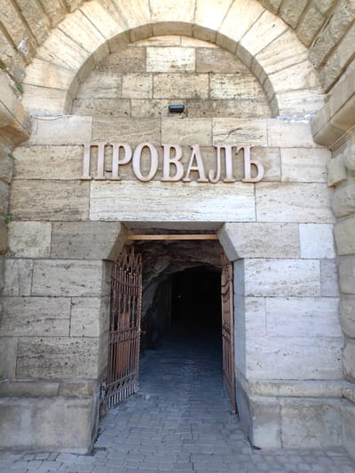 The image shows an entrance to a cave or tunnel in Goryachevodsky, Russia. Above the entrance, large wooden letters spell out "ПРОВАЛЬ" which translates to "Collapse" or "Sinkhole" in Russian. The entrance is framed by a stone archway and is partially obscured by a rusty metal gate, which is open. The ground in front of the entrance is paved with cobblestones. The overall scene suggests a historical or geological site that is accessible to the public.
