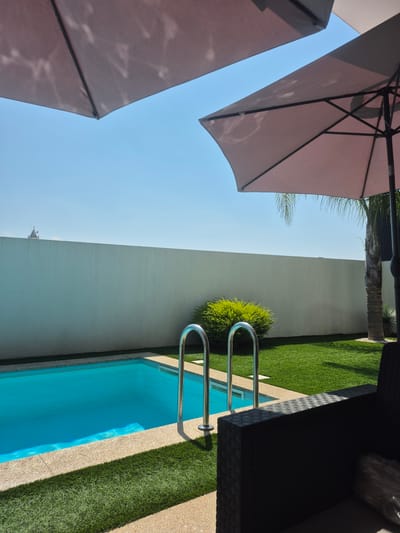 This media file captures a sunny day outdoors, likely in Felgueiras, Portugal. The scene features a swimming pool with vibrant blue water, surrounded by artificial turf and a beige concrete deck. A silver metal ladder with two steps enters the pool, indicating it's ready for use.
In the foreground, parts of two large, rose-colored patio umbrellas are visible, angled upwards and casting dappled shadows. Their structure suggests they are providing shade. To the right, a dark, woven-textured piece of furniture, possibly a sofa or lounger, is partially visible, also casting a shadow.
The background is dominated by a clean, off-white wall that curves slightly. Beyond the wall, hints of greenery, including palm fronds and a small, dense green shrub near the pool ladder, suggest a garden or landscaped area. The sky is a clear, bright blue, with no clouds visible. In the far distance, a small architectural element with a red roof and spires can be seen peeking over the wall, adding a touch of detail to the horizon.
The lighting suggests it is daytime, likely mid-morning to afternoon, given the strong sunlight and defined shadows. The overall impression is one of a relaxed, vacation-like atmosphere, ideal for swimming and outdoor leisure. No people are visible in the frame. There is no visible text in the image.