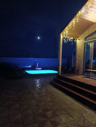 The image depicts a nighttime scene in the city of Lenina, Russia, featuring a modern wooden house with illuminated porch lights. To the left, a swimming pool glows with an aquamarine light, and a pink inflatable flamingo float is visible in the water. The yard is paved with dark tiles, wet from recent rain, reflecting the lights. A corrugated metal fence encloses the property, and a distant power line tower is silhouetted against the dark blue sky, where a sliver of the moon is visible. The scene suggests a private residential setting.
