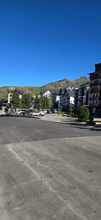 The image depicts a sunlit scene in Sallent de Gállego, Spain, with a clear blue sky overhead. In the foreground, a section of dark asphalt pavement is visible, showing some wear and tear with scattered white specks and tire marks. Beyond the pavement, a gently curving road leads towards a row of multi-story buildings characteristic of a mountain resort town. These buildings, with their dark tiled roofs and white or beige facades, are nestled against a backdrop of rolling, green hills that transition into rocky, arid mountain peaks further in the distance.

Several vehicles are parked along the road in front of the buildings, including white SUVs and a dark-colored van. Lush green coniferous trees are interspersed throughout the scene, adding to the natural beauty of the location. Some of these trees are planted in neat rows, suggesting landscaping. Streetlights and decorative planters are also present, indicating a well-maintained urban or semi-urban environment. The architecture of the buildings suggests they are likely hotels or apartment complexes, catering to tourists. The overall impression is one of a peaceful mountain village on a clear, bright day.