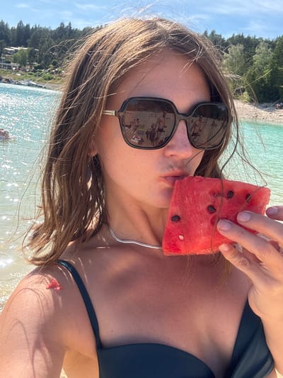 An outdoor, close-up, self-portrait photograph captures a woman at the Lac de Vouglans, a reservoir in Maisod, France, on a sunny day. The woman, positioned in the foreground, is looking towards the camera and holding a slice of watermelon to her lips. She has brown hair that is blowing in the wind, and she is wearing large, dark-rimmed sunglasses that reflect a scene of people picnicking on a beach. She is also wearing a dark green bikini top. A silver necklace is visible around her neck. In the background, the bright turquoise water of the lake is visible, with trees and a clear blue sky above. A small piece of pink material is on her left shoulder.