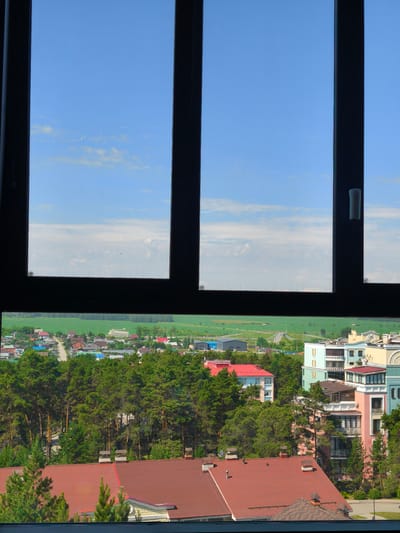 This is a photograph taken from a window looking out over a town and surrounding landscape. The view is framed by a dark window frame with vertical and horizontal mullions, dividing the scene into four panes.

In the upper section of the photograph, a bright blue sky dominates, with a few scattered, wispy white clouds. The sunlight suggests it is daytime, likely mid-morning or afternoon. The quality of light and the clear sky indicate fair weather.

The lower section of the photograph reveals a residential area with a variety of buildings. Closer to the foreground, a large building with a reddish-brown tiled roof is prominent. Several other buildings with red, blue, and gray roofs are visible, interspersed with lush green pine trees. Beyond this, the town extends further, with more houses and larger buildings, including multi-story structures with balconies and colorful facades. A road winds through the town. In the distance, a vast, flat green field stretches out, meeting the horizon under the blue sky. The overall impression is of a peaceful, suburban or semi-rural setting in Russia, possibly Shigaevo, given the provided location context. There are no people visible in the image, and the focus is on the architecture and the natural landscape. The image appears to be a static, observational shot, without any specific activity or event taking place.