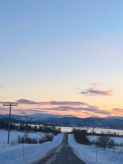 The scene depicts a winter landscape in Grasmyr, Norway, at either sunrise or sunset. A snow-covered, dark asphalt road occupies the lower center, sloping downwards and curving slightly to the right, flanked by deep snowdrifts. Two red/orange reflective poles mark the right edge of the road. To the left, a tall utility pole with several crossarms and electrical wires stands in the snow. The midground features snow-covered fields with sparse, leafless deciduous trees and bushes. Beyond these fields lies a body of water, likely a fjord or bay, reflecting the sky. On the distant right, near the water, a dark, small structure or building is visible. The background consists of a range of snow-capped mountains, appearing dark blue-purple, silhouetted against the horizon. The sky transitions from light blue at the zenith to hues of orange, pink, and purple clouds along the mountainous horizon. No people, animals, or specific actions are observed.