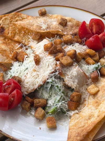 This is a close-up, overhead shot of a Caesar salad on a white plate. The salad features grilled chicken strips, croutons, shredded Parmesan cheese, and sliced cherry tomatoes. Large, golden-brown tortilla chips are layered around the salad. The plate is set on a wooden surface, likely a table. The background is slightly blurred, suggesting an outdoor or well-lit indoor setting. There are no people visible in the image. This scene is located in Byurakan, Armenia.