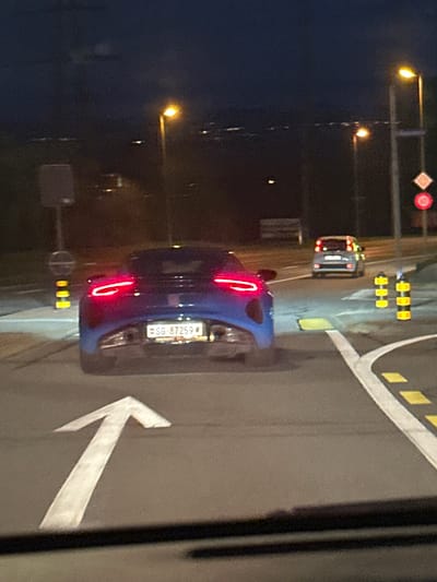 The image captures a nighttime street scene in Volketswil, Switzerland. A dark blue sports car, likely an Alpine A110, is the central subject, viewed from the rear on an asphalt road. Its two prominent red LED taillights are illuminated. A Swiss license plate, "SG 87259", is affixed to the rear bumper.
The vehicle occupies a lane marked by a faded white arrow on the pavement indicating forward travel. White curved lines and yellow rectangular road markings delineate a right turn lane or intersection. Two yellow and black striped traffic bollards are positioned to the right of the vehicle.
Further down the road, a silver-grey compact car is visible from the rear with illuminated taillights. The environment is illuminated by multiple yellow-orange streetlights. Infrastructure includes a grey pole with an indistinct square sign and a yellow-black striped pole on the left. On the right, traffic signs are visible on a pole: a white diamond sign, a red circular 'no entry' sign, and a red circular '50 km/h' speed limit sign. Distant city lights are visible on the horizon beneath a dark sky. No individuals are discernible.